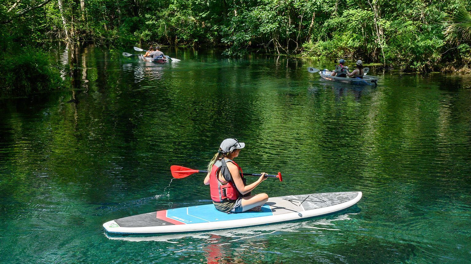 Woman paddles a stand-up paddleboard on clear, blue water. Lush green trees surround the waterway.