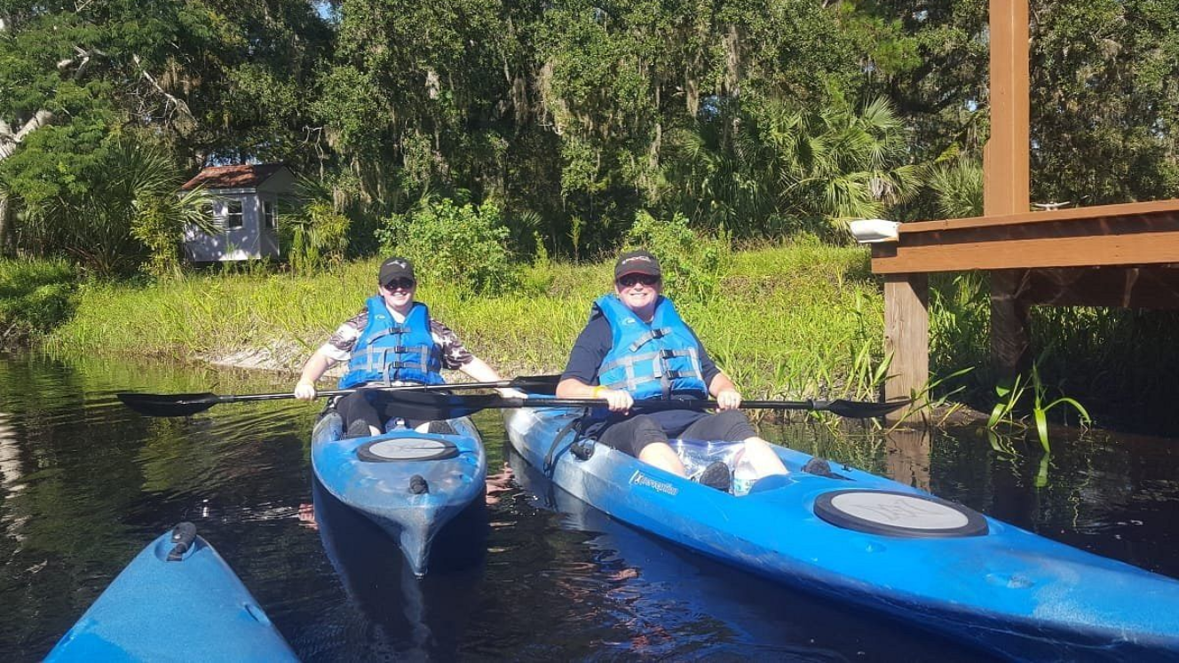 Two people in blue kayaks paddling on a dark waterway near greenery and a wooden dock.