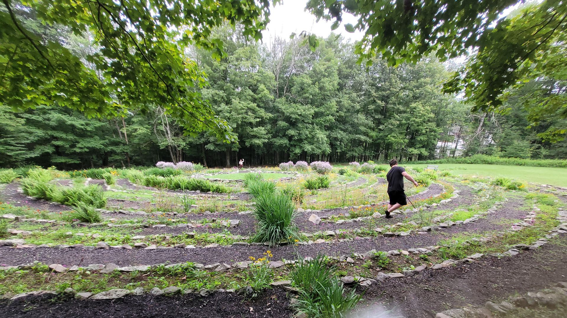 A person walks a circular garden labyrinth with stone pathways, surrounded by plants and trees.