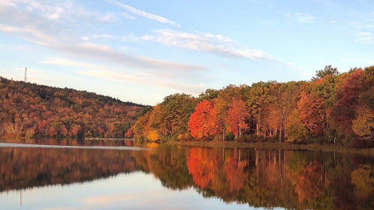 Autumn trees with red, orange, and yellow leaves reflected in calm water. Blue sky and soft clouds.