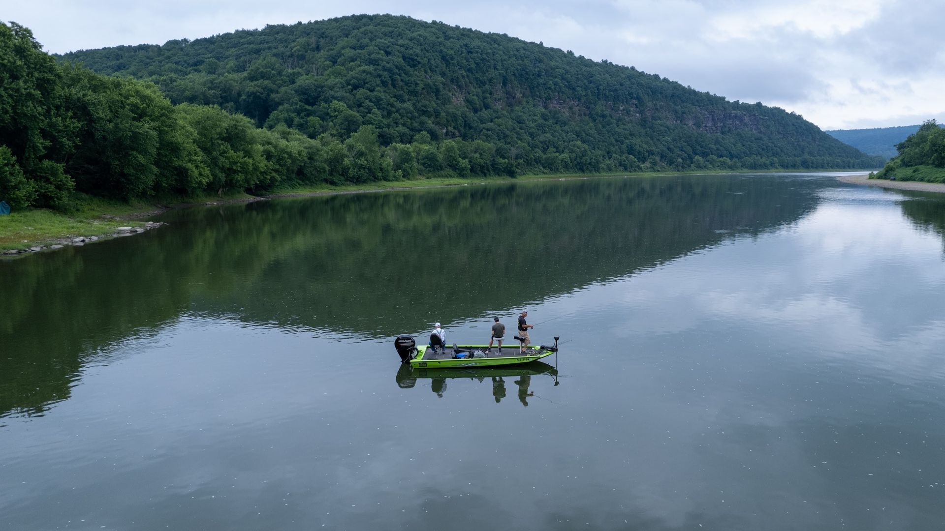 Boat on calm water with three people fishing, mountain in the background. Green boat, gray water, overcast sky.