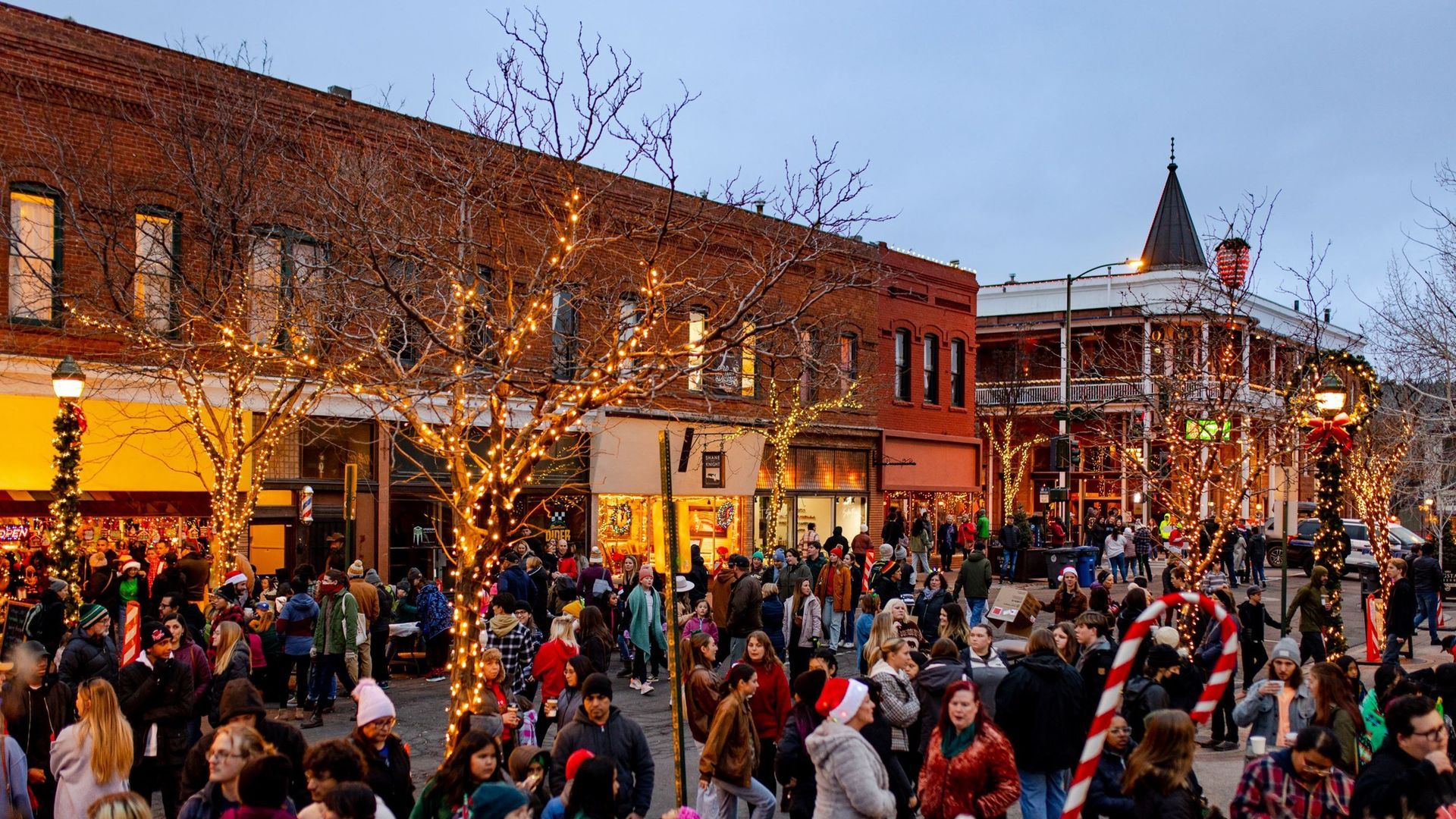 A festive downtown street scene at dusk featuring crowds of people, holiday lights on trees, and red brick storefronts.