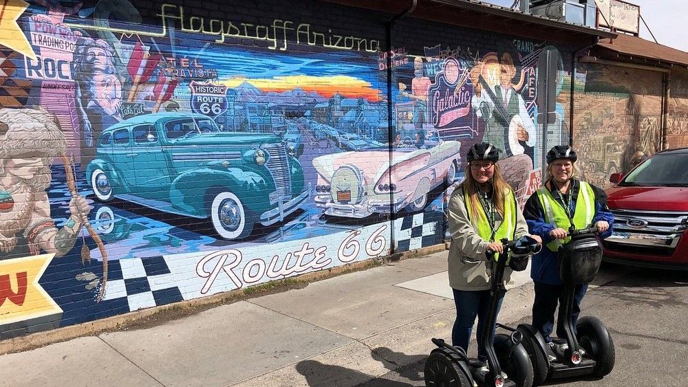 Two people on Segways stand before a colorful Route 66 mural in Flagstaff, Arizona, featuring classic cars.