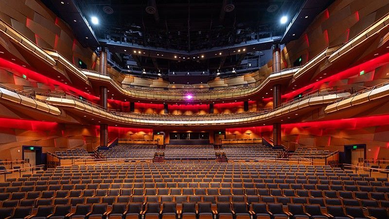 Interior of a grand theater with rows of seats facing a stage. Red lighting accents the balconies and walls.