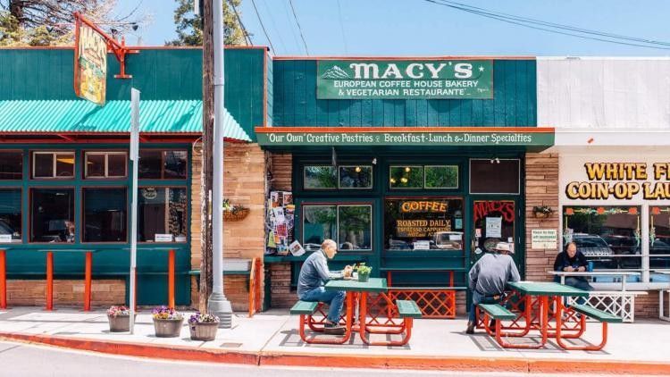 Two people sit at outdoor tables in front of Macy's, a green-fronted restaurant with a bakery, on a sunny day.