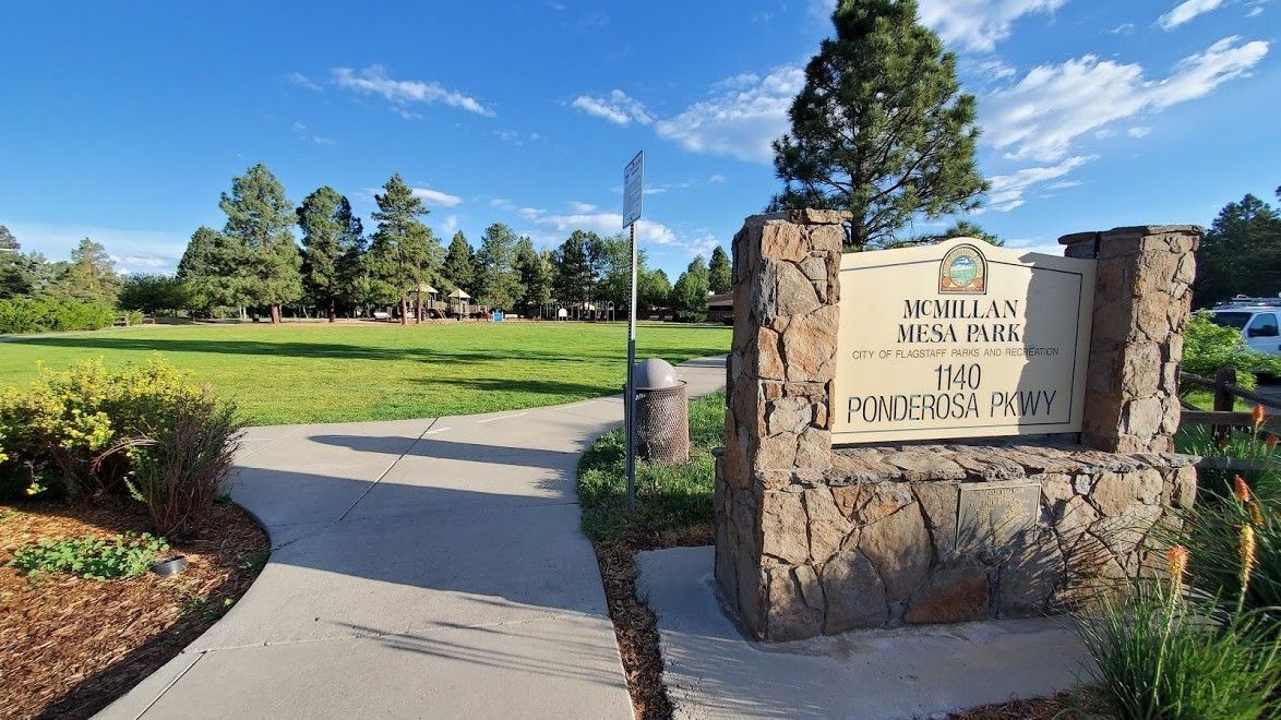 Park entrance with stone sign, paved path, green grass, trees, and a blue sky.