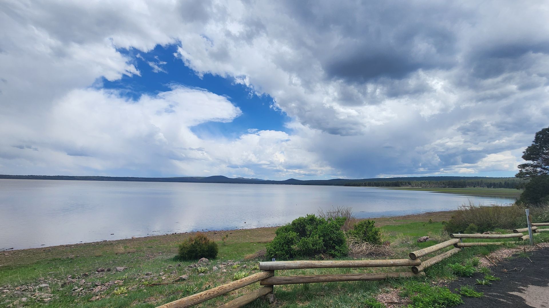 Lake with overcast sky, dark clouds, and grassy shoreline with wooden fence.
