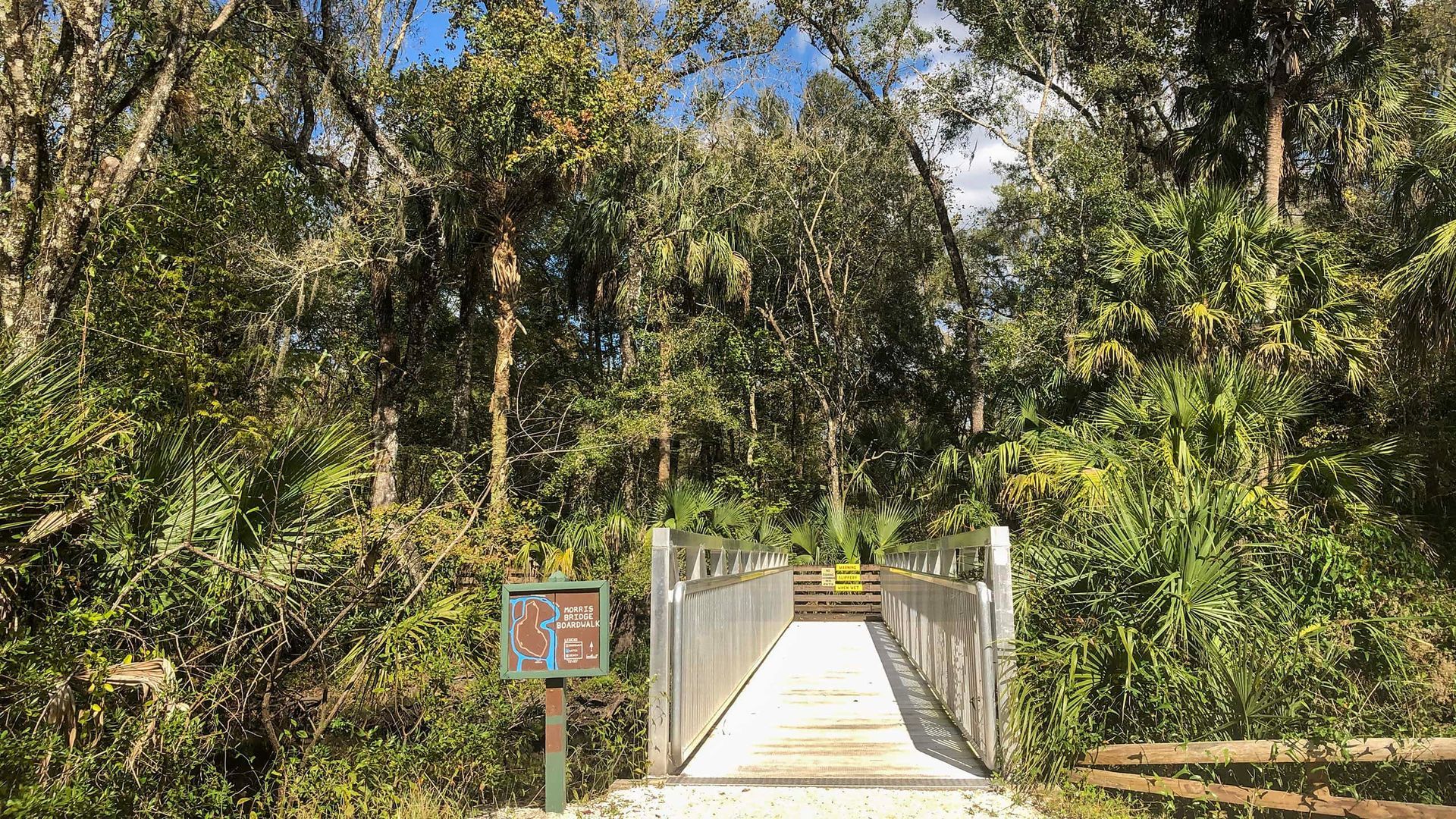 Wooden bridge over a shaded trail, surrounded by lush green foliage and trees under a partly cloudy sky.