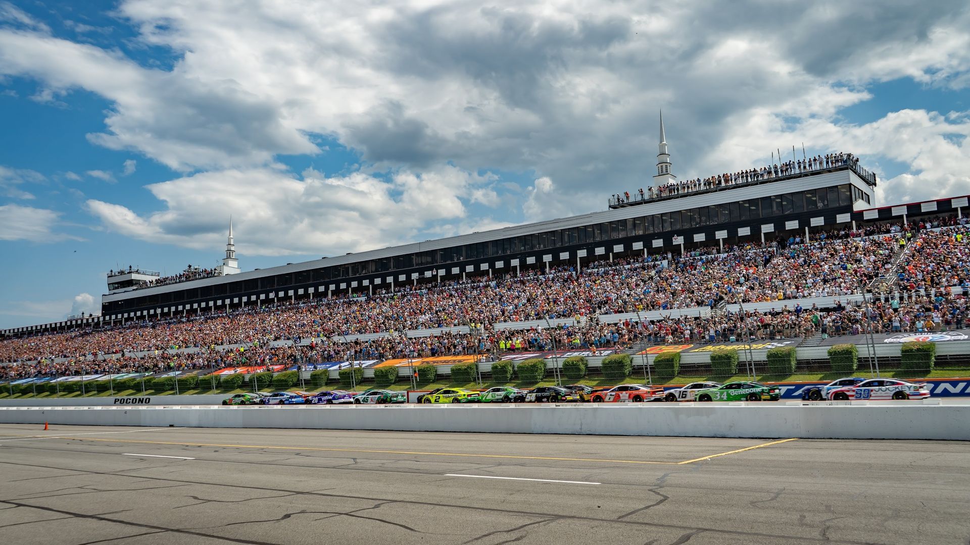 A line of race cars driving on a track in front of a crowded stadium grandstand under a blue sky with white clouds.