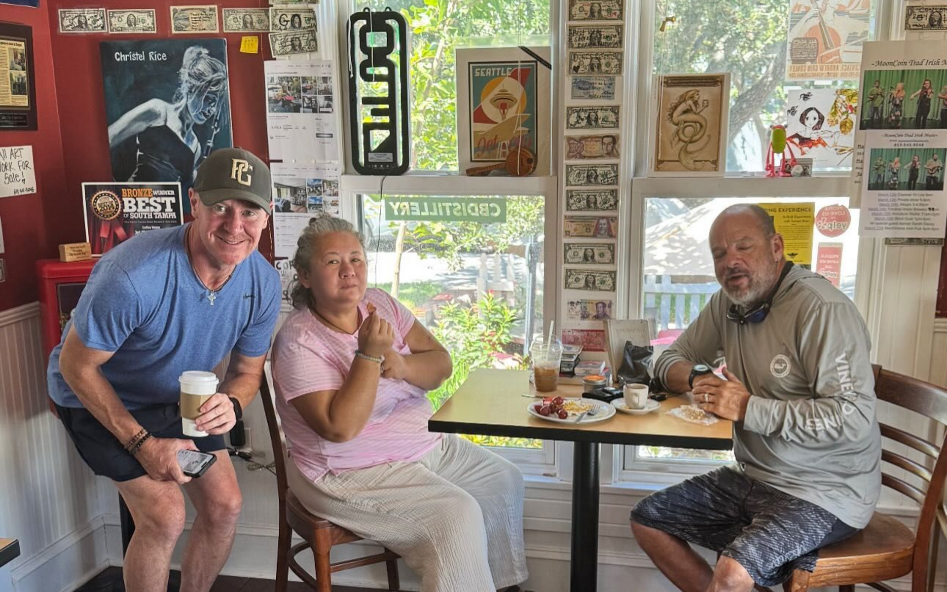 Three people seated and standing in a cafe with wood floors. A person is holding a coffee cup, another is sitting with hands clasped.