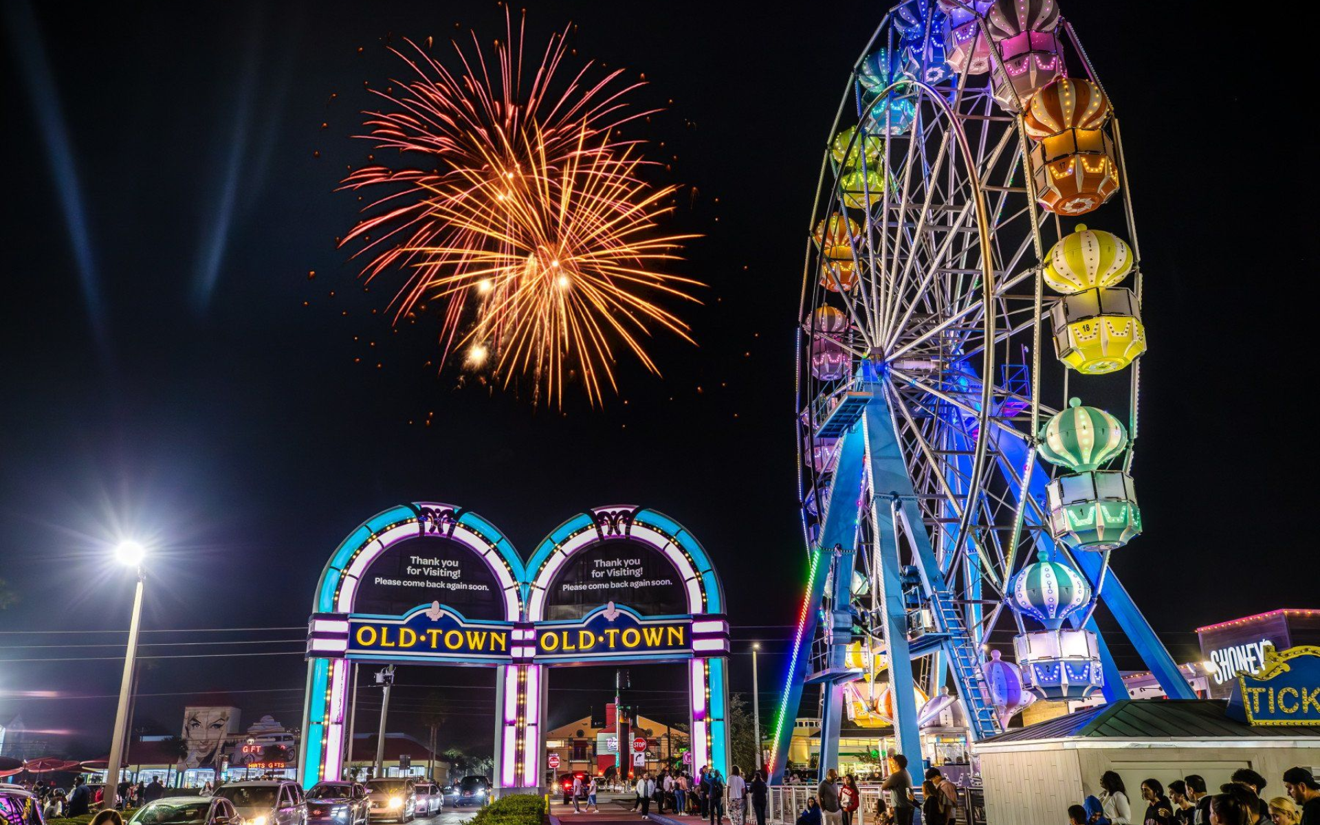 Fireworks burst over Old Town amusement park entrance and a lit Ferris wheel at night.