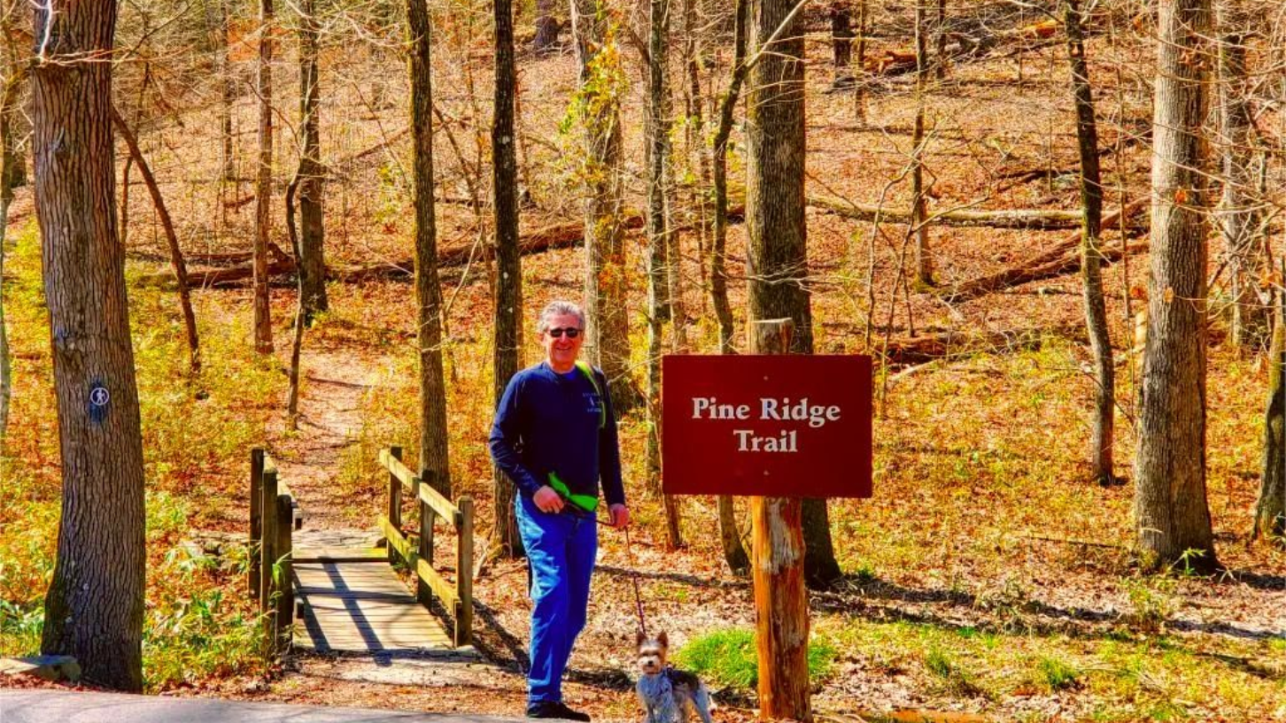 Man standing at Pine Ridge Trail sign with a dog; autumn forest setting.