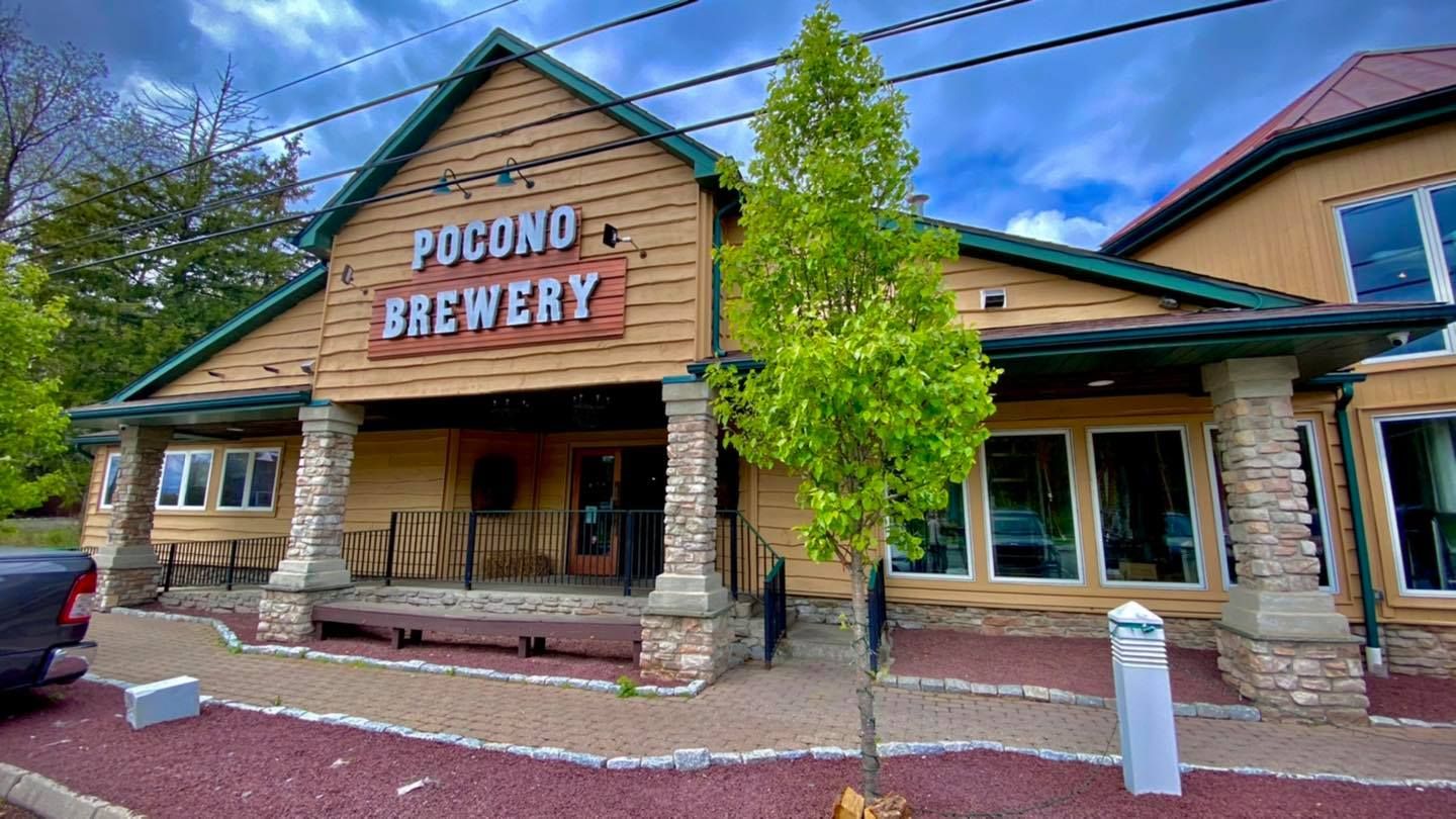 The Pocono Brewery, a rustic building with wooden siding, stone pillars, and a green roof, under a cloudy blue sky.