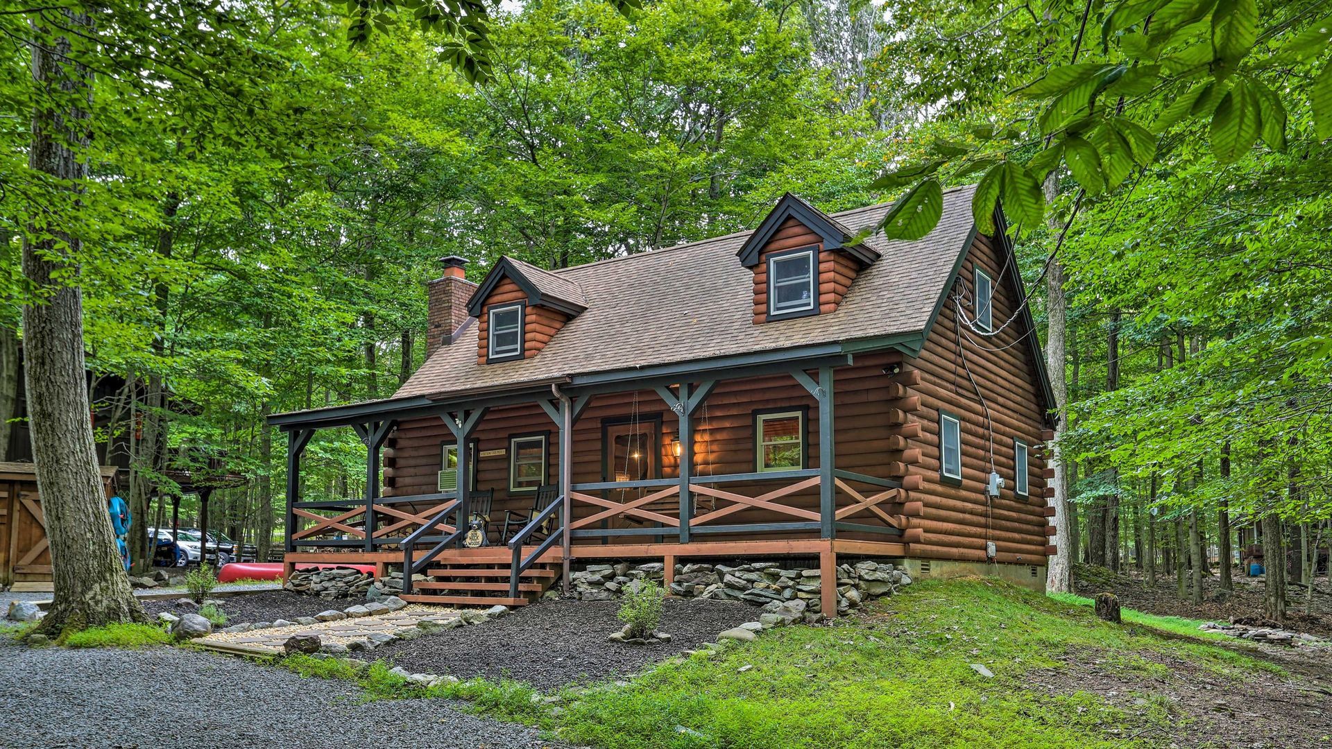 Log cabin nestled in green trees, with a porch and two dormers.