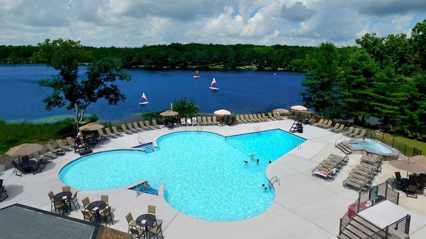 Pool and lake view: freeform pool, sunbathers, sailboats, trees, blue water, cloudy sky.