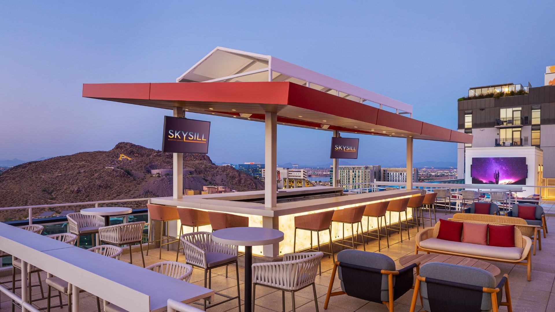 Rooftop bar with illuminated bar, seating, and cityscape background at dusk.