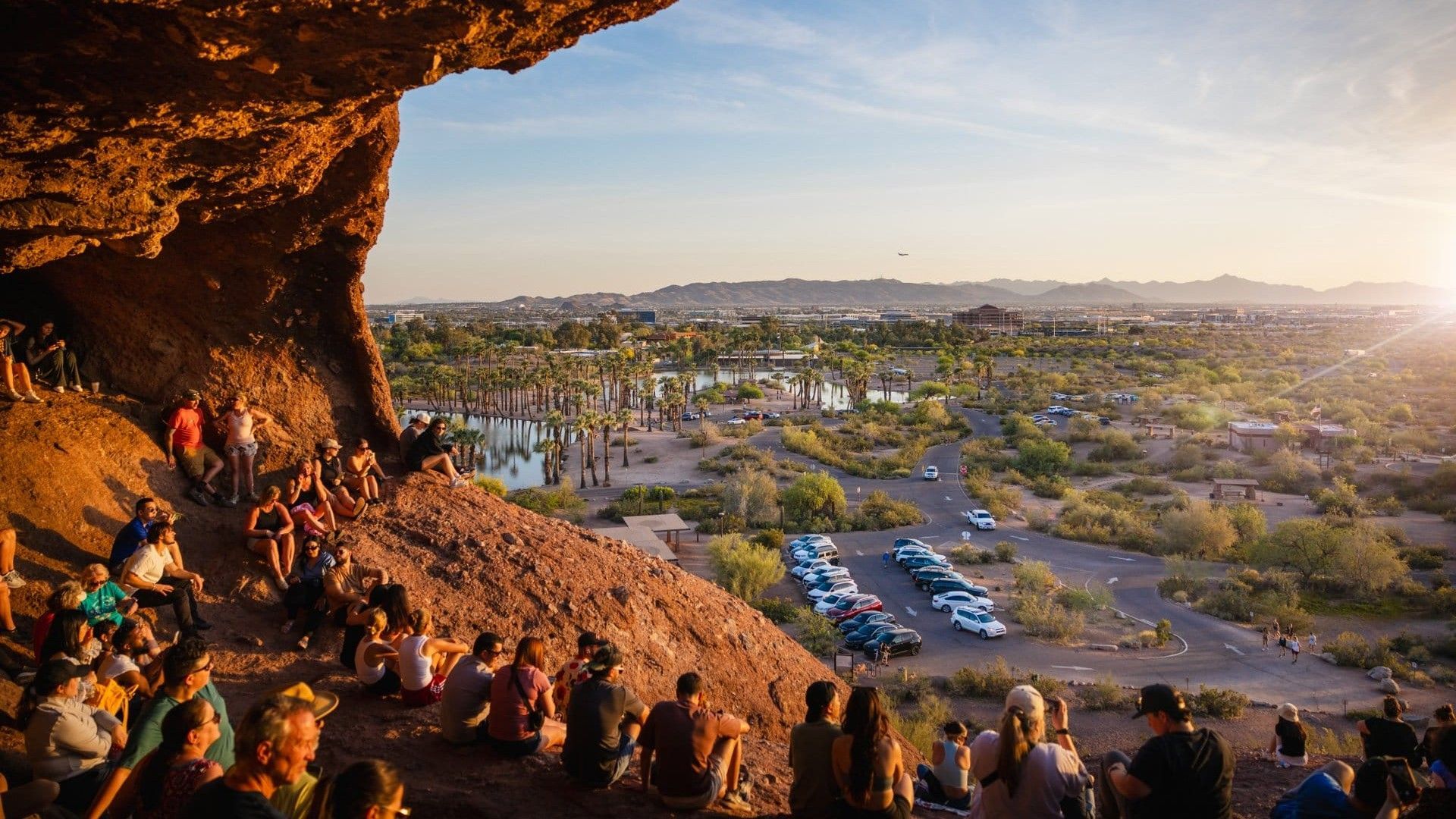 People sit on a rocky hillside overlooking a desert landscape with a lake and palm trees.