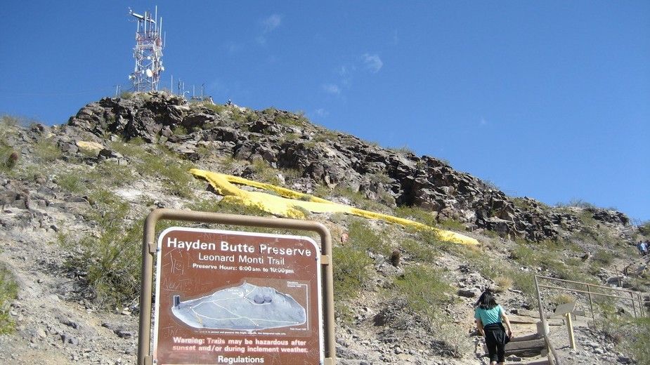 Sign for Houston Mountain Trail with a person hiking upward towards the mountain's peak with a cell tower, sunny day.