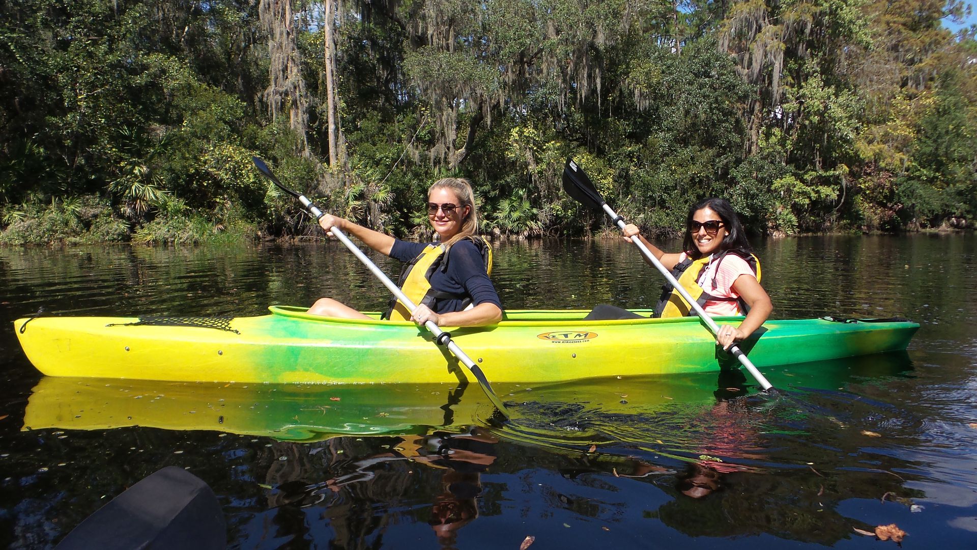 Two people kayaking on a calm river. Yellow and green kayak, sunny day, wooded background.