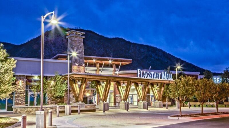 Exterior view of Flagstaff Mall at twilight, featuring a wooden canopy entrance, stone pillars, and mountain backdrop.