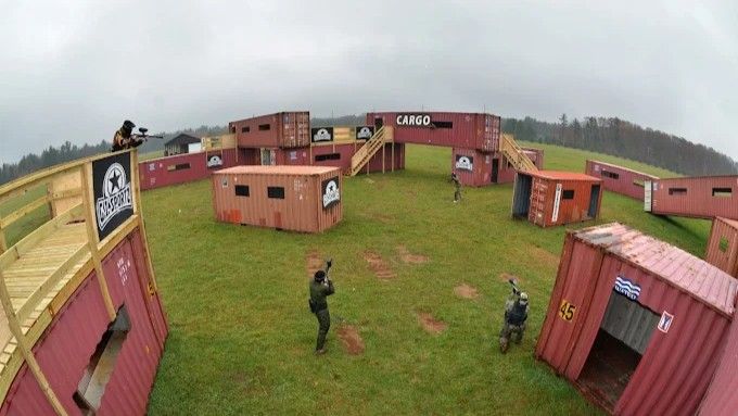 Paintball game in outdoor field. Players shooting around red shipping containers and wooden structures.