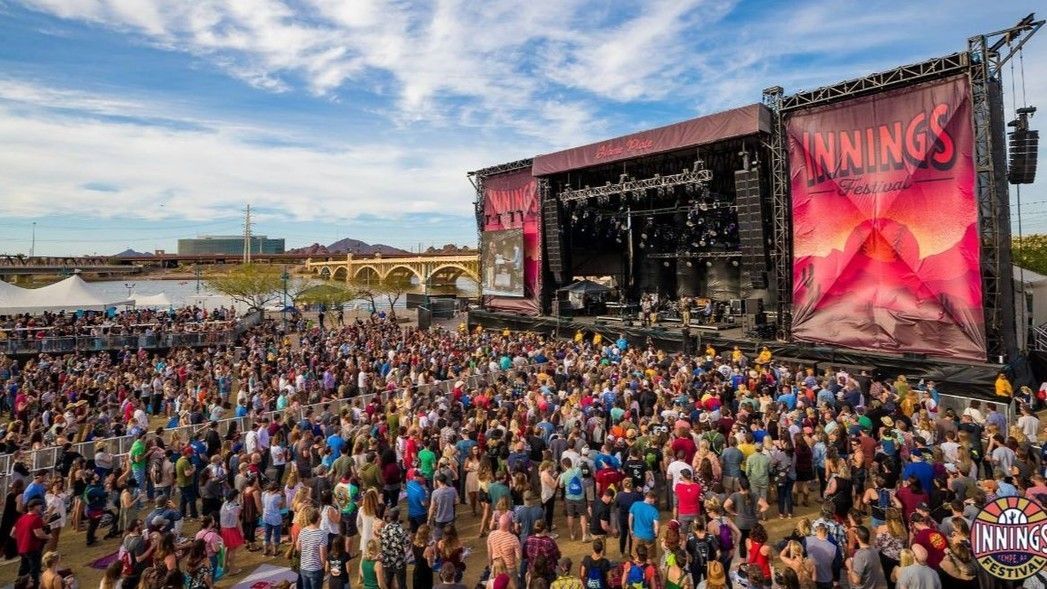 Large outdoor music festival with crowd in front of stage under a bright blue sky.