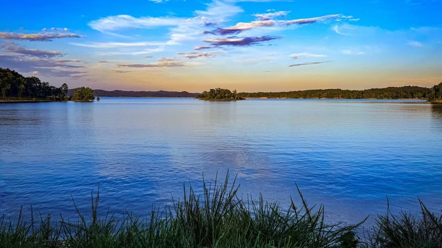 Calm lake under a blue and orange sunset sky, small island and green foliage in foreground.