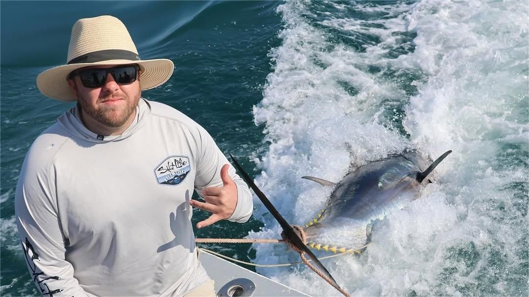 Man on boat with a large fish jumping in the ocean; he's giving a