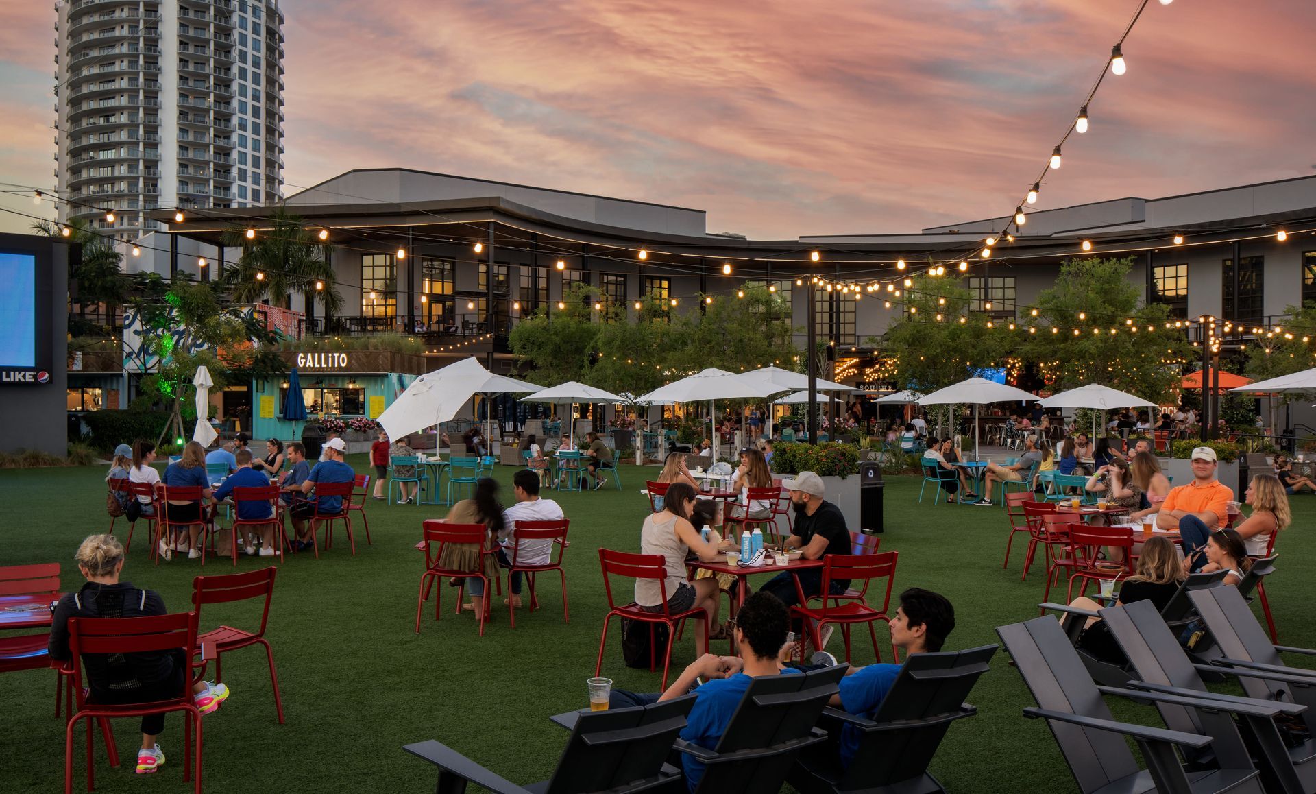 People seated at tables on a lawn, watching a screen at dusk, string lights overhead.