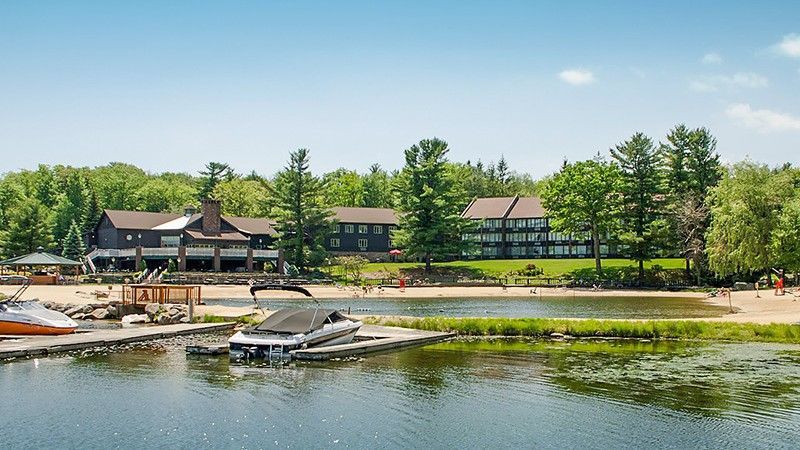 Lakeside resort with dock, beach, and dark buildings under a sunny sky and green trees.
