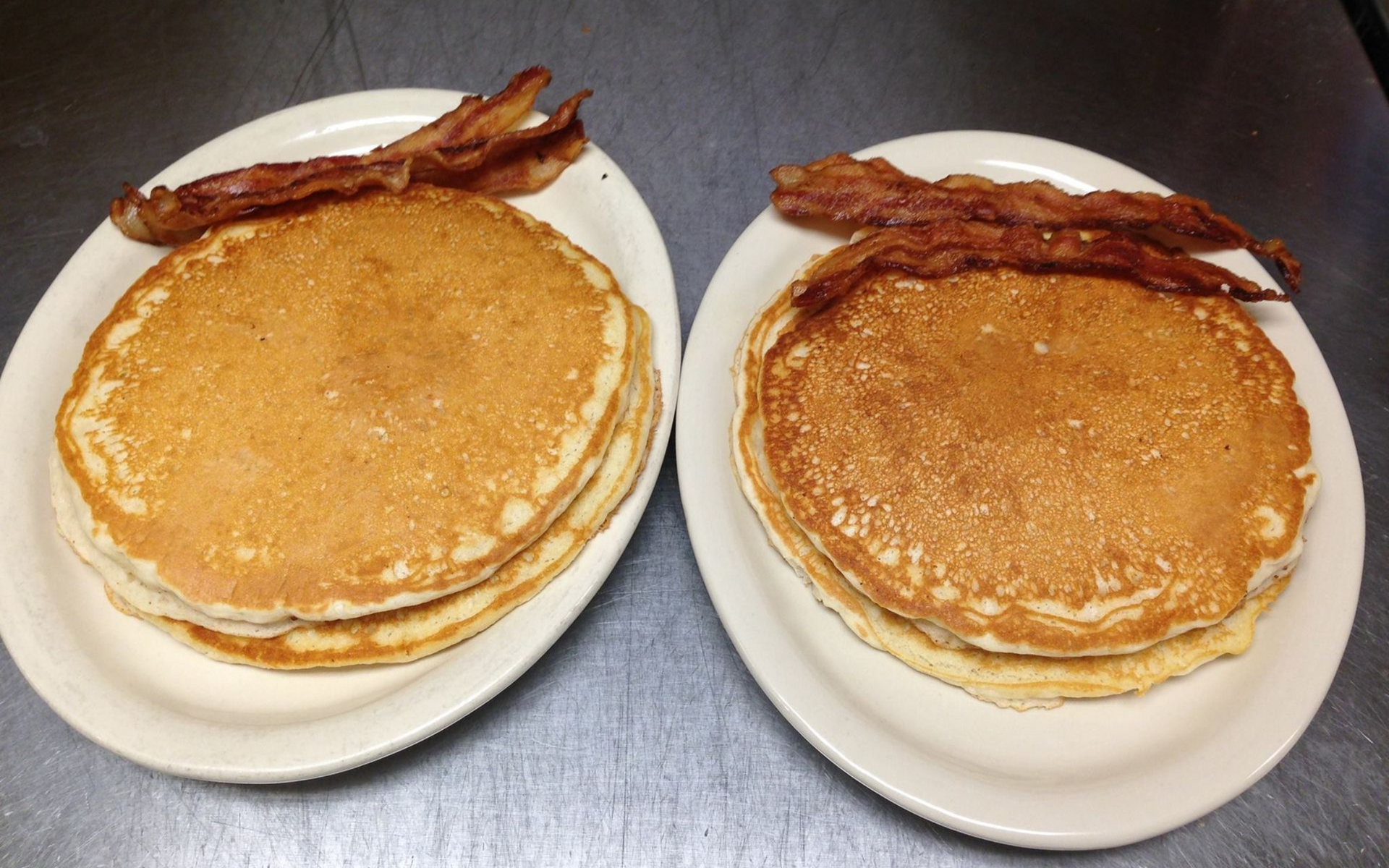 Two plates of pancakes with bacon on a stainless steel surface.