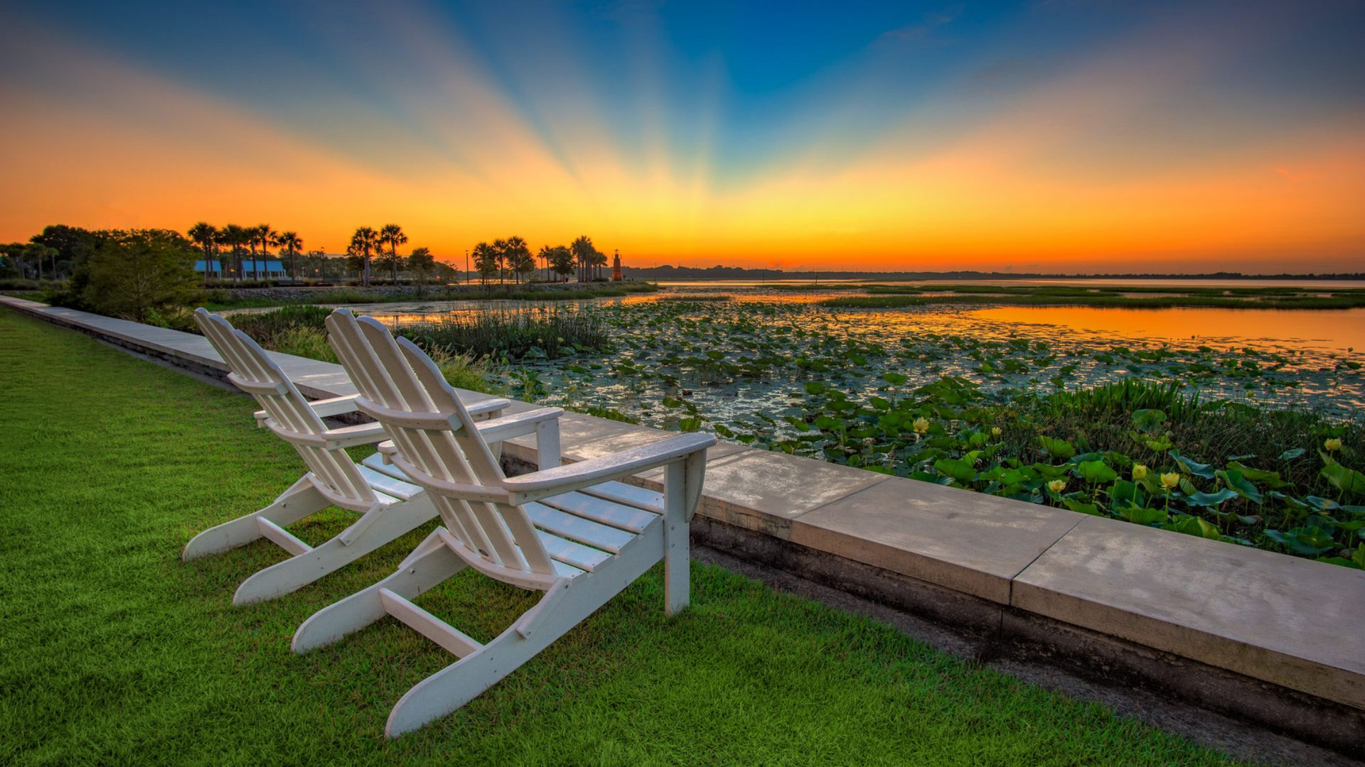 Two white Adirondack chairs on green grass overlook a marsh at sunset, with orange and blue sky.