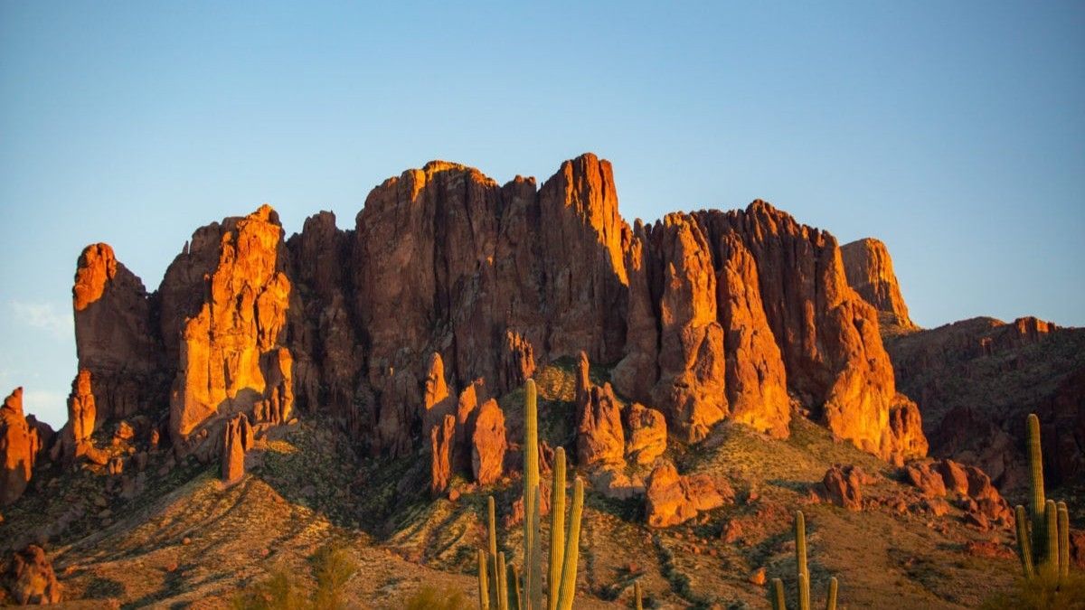 Rugged, golden-lit mountain range with saguaro cacti under a clear blue sky.