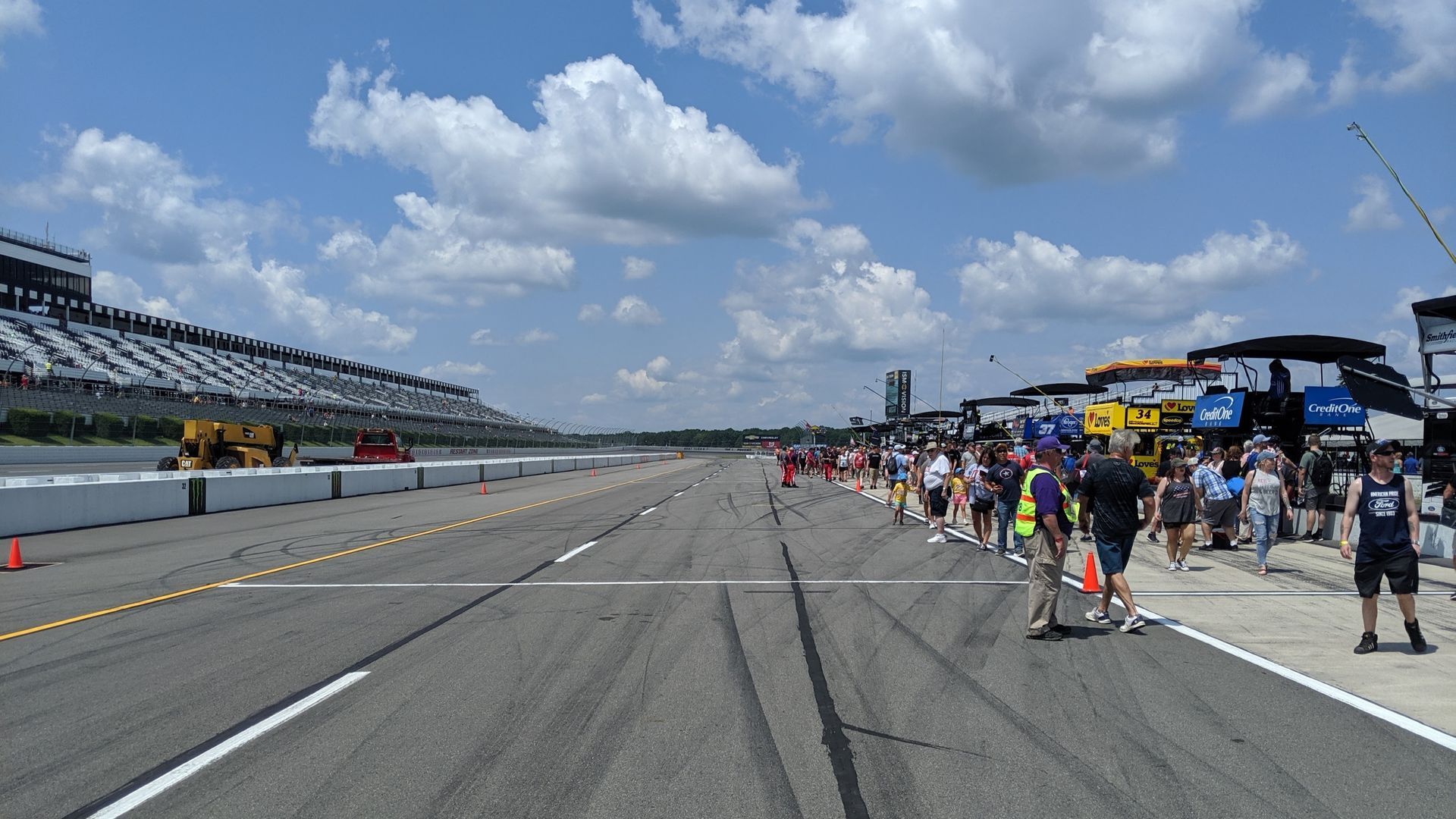 A view of an empty race track with people standing along the pit lane under a blue, cloudy sky.