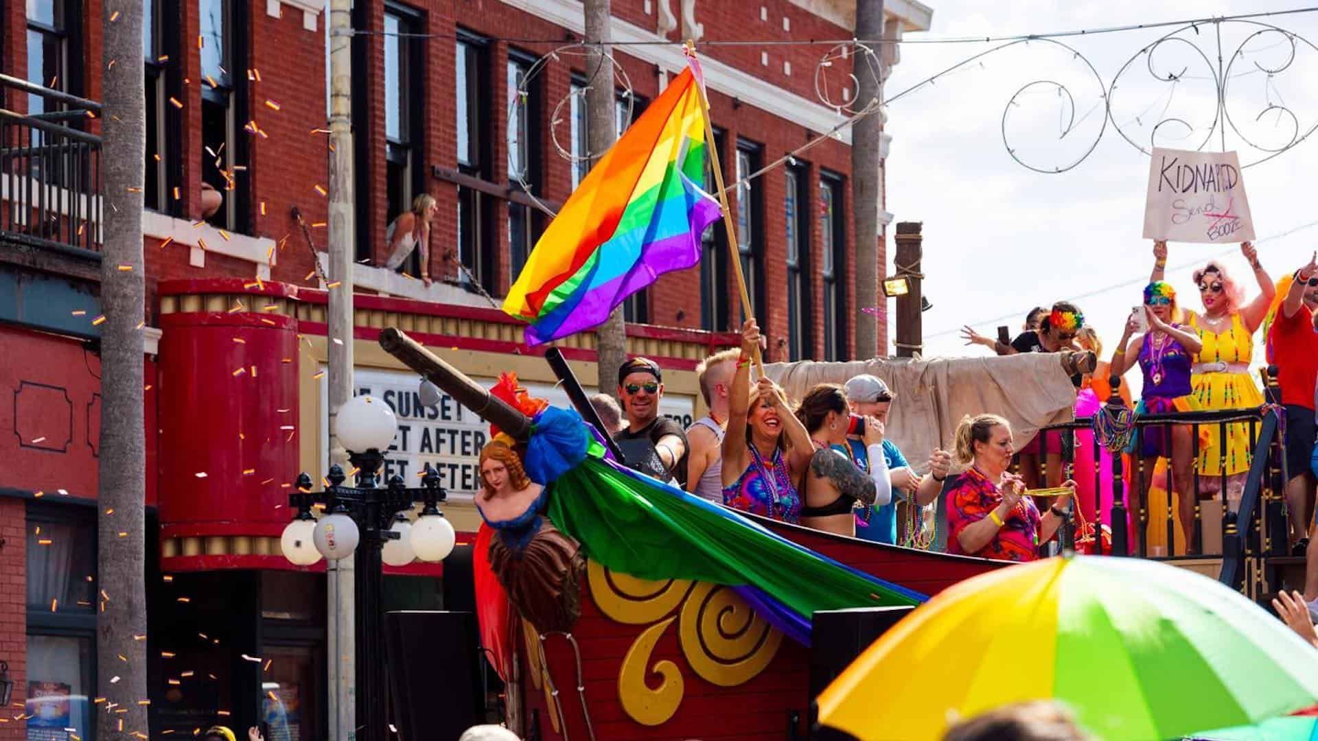 People celebrating on a float at a Pride parade, waving a rainbow flag. Buildings with onlookers.