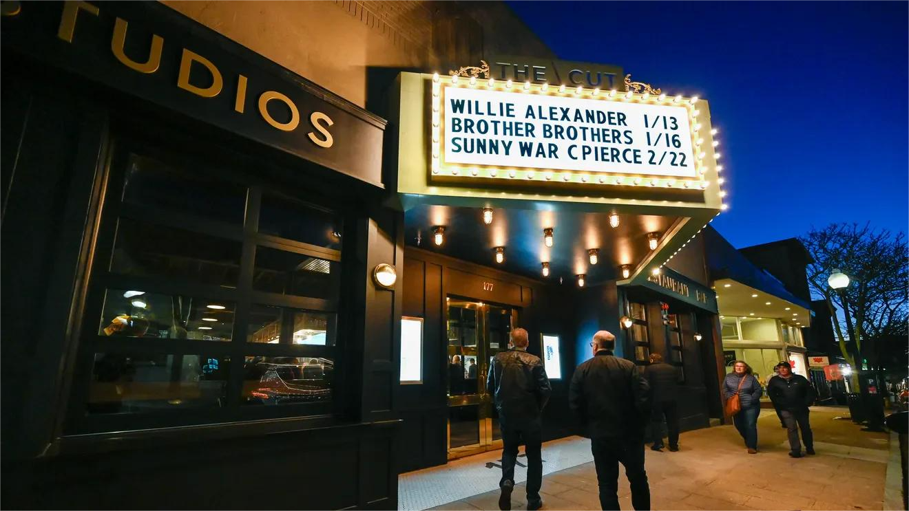 Theater facade with illuminated marquee displaying artist names and dates, people outside at dusk.