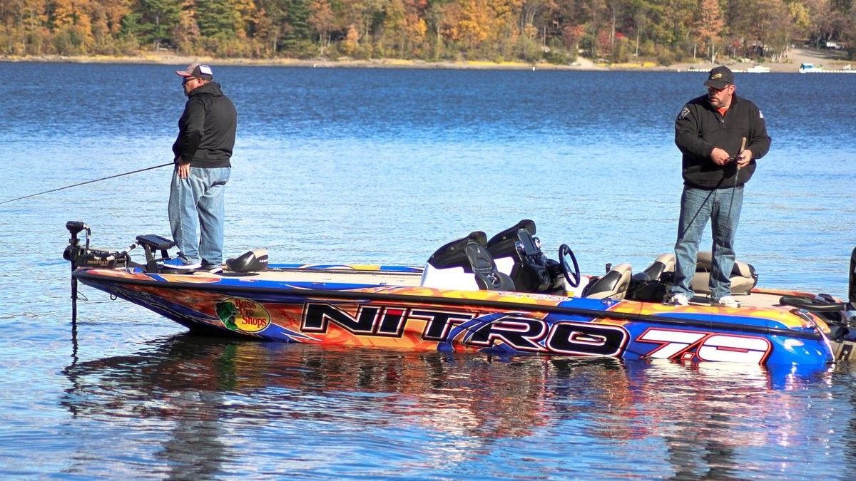 Two people fishing from a colorful Nitro bass boat on a lake with trees in the background.
