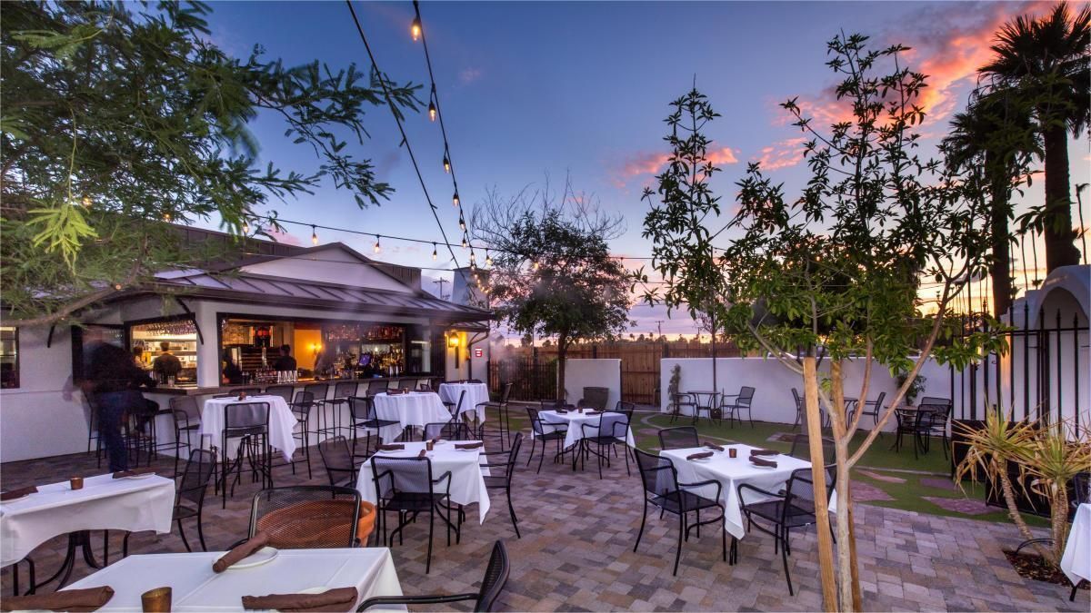Outdoor restaurant patio at dusk with tables, chairs, bar, and string lights under a colorful sky.