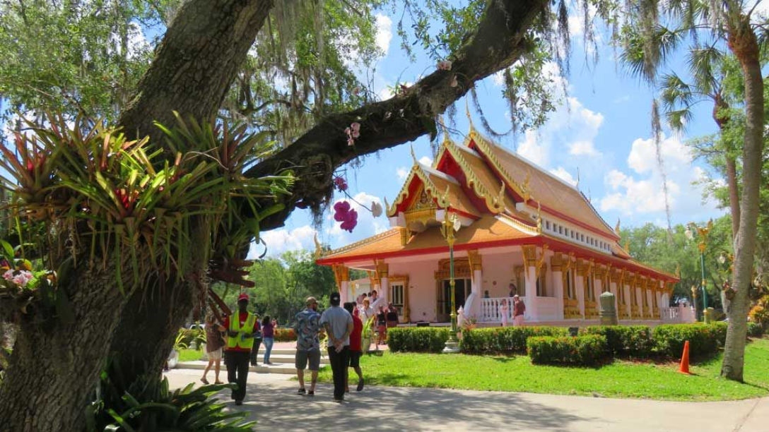 Thai Buddhist temple with gold roof, pink walls, people walking toward it, sunny day.
