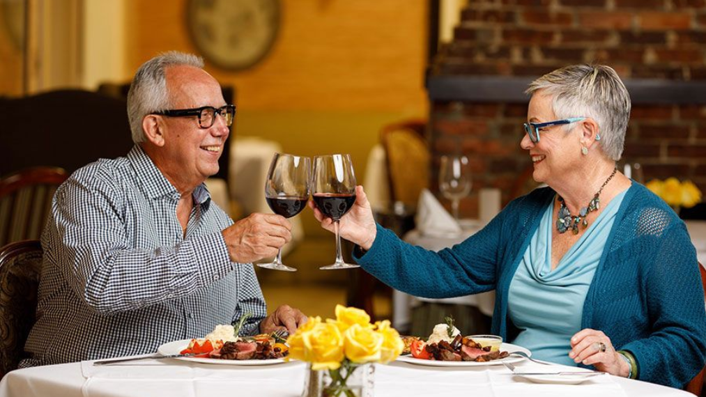 Senior couple toasting wine glasses at a restaurant table. They are smiling, with food and flowers present.