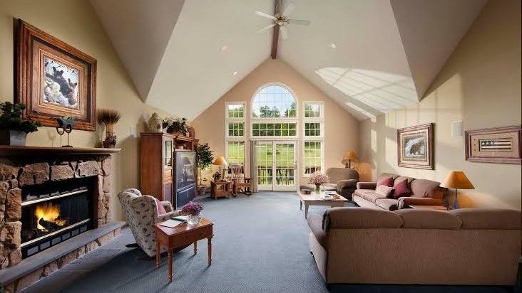 Cozy living room with fireplace, vaulted ceiling, large windows overlooking a green lawn, and brown furniture.