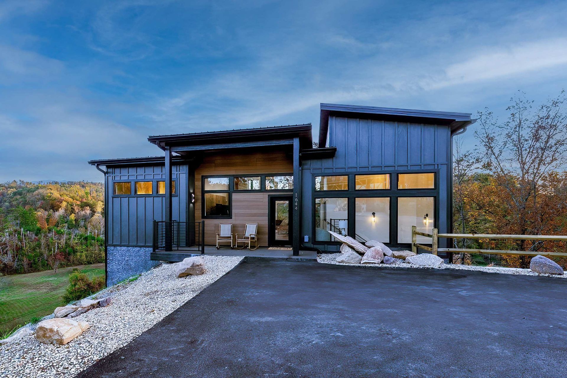Modern dark-blue house at dusk with warm interior lights, paved driveway, and a rocky landscaped front yard