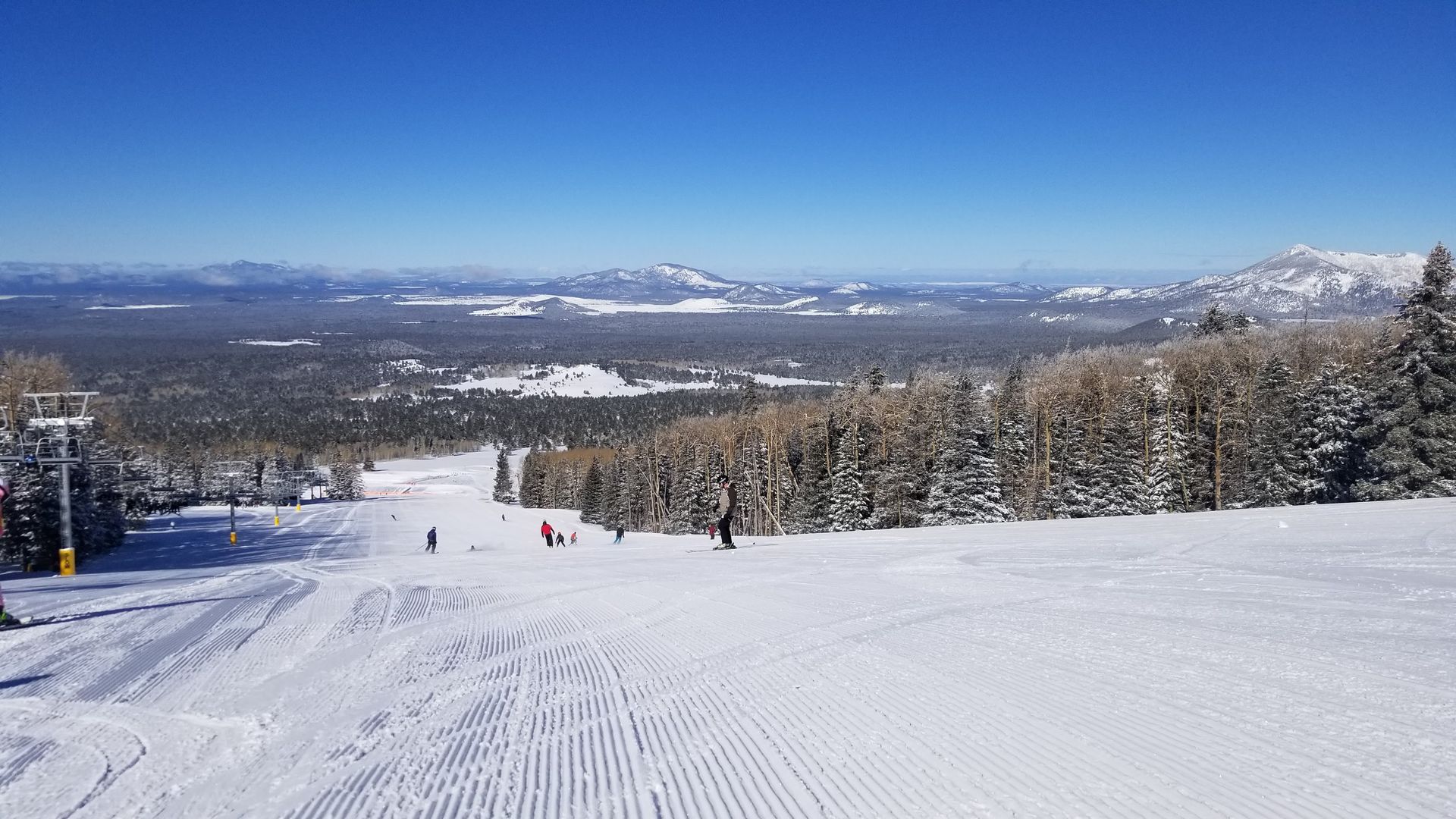 Snowy ski slope with skiers and snow-covered trees under a clear, blue sky.