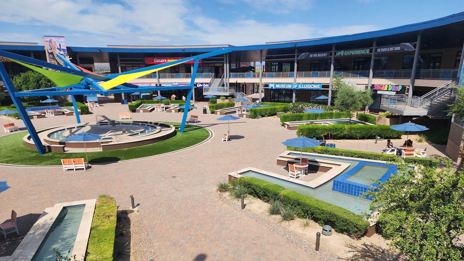 Outdoor shopping plaza with blue canopies, water features, seating, and a blue sky.
