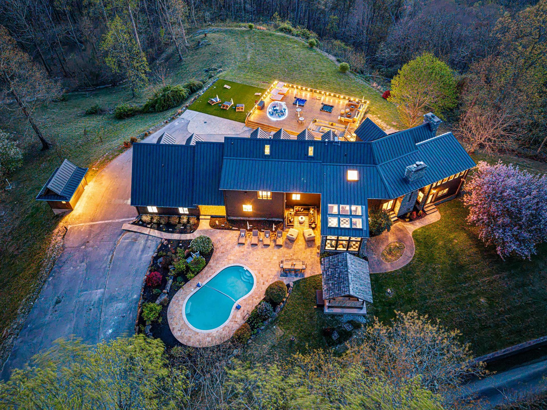 Aerial view of a large house at dusk with lit patio, pool, and surrounding trees.