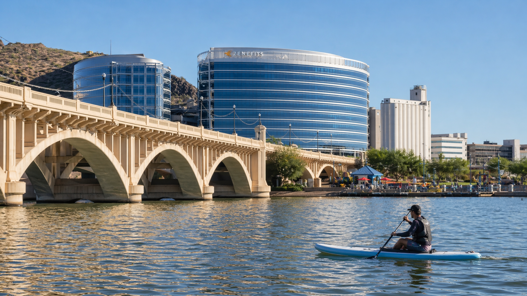 Stone bridge over water with modern buildings behind and a person kayaking in the foreground