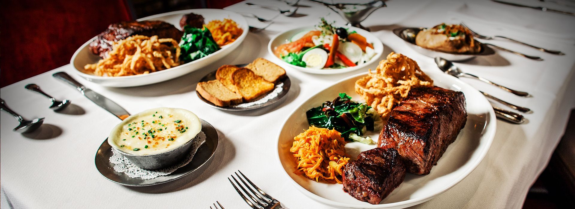 Platters of food on a white tablecloth; featuring cooked vegetables, meat, and bread.