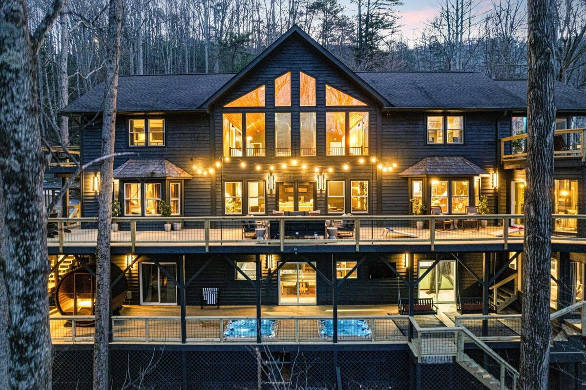 Large illuminated lakeside lodge at dusk, with warm lights glowing through tall windows and a wooden deck.