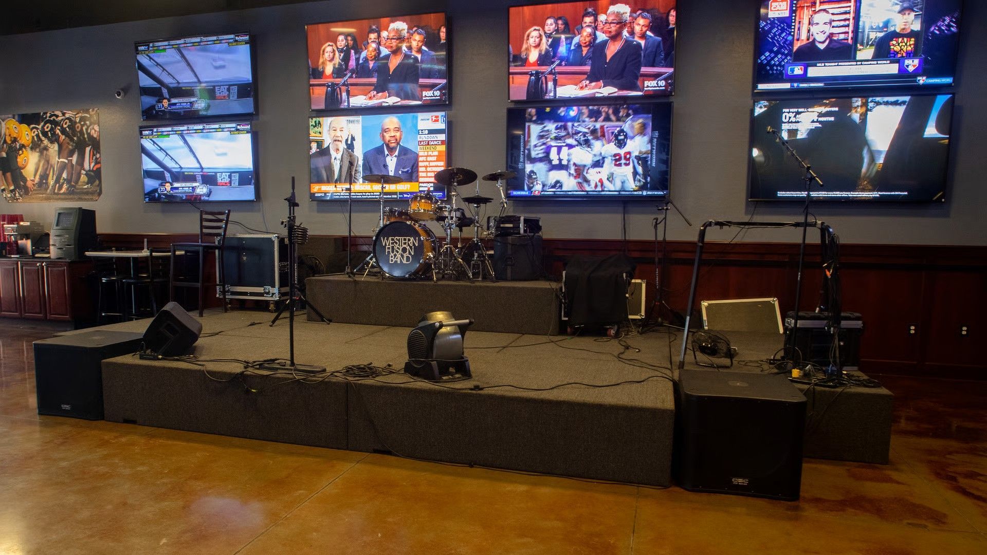 A stage with a drum set, microphones, and speakers. Multiple TVs show various live feeds. Brown flooring.