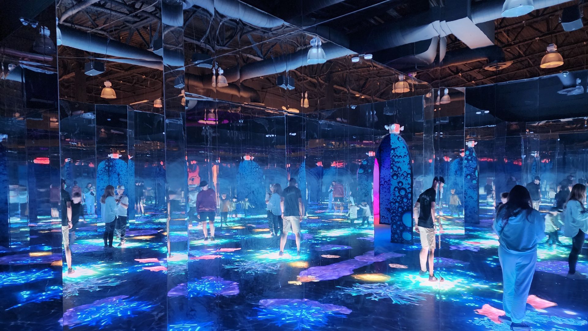 Mirrored room with people walking. Blue and purple lights on the floor and walls.